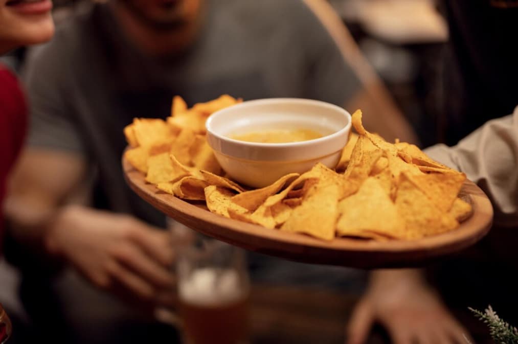 A wooden tray with nachos and a bowl of dip shared by people